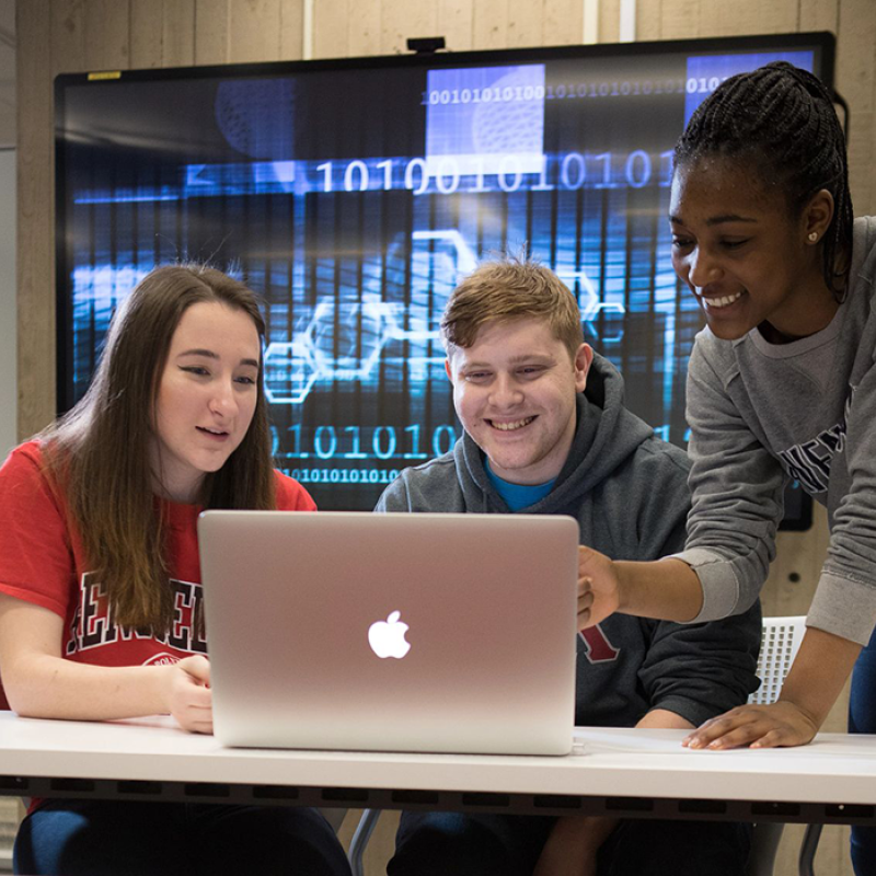 Three students work on a laptop in front a screen with computer code